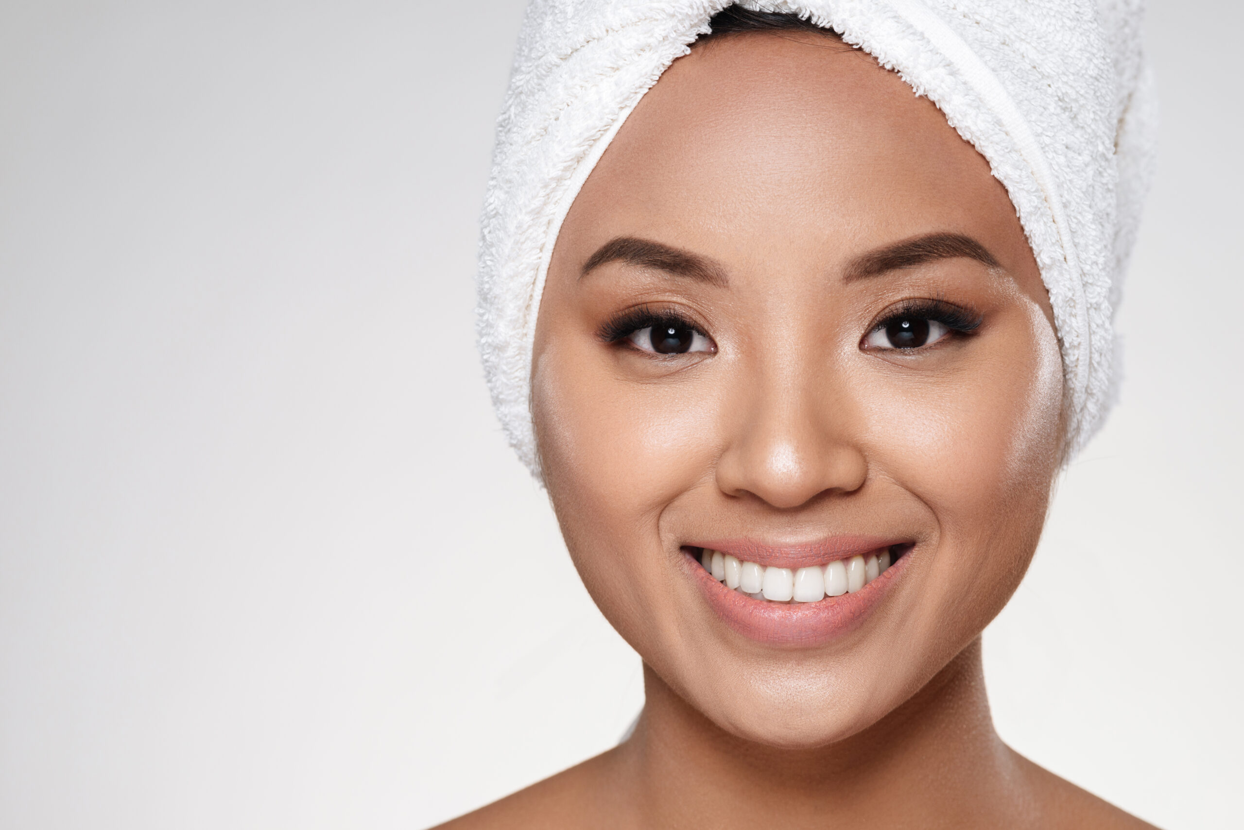 Attractive asian half-naked lady without make up with towel on head smiling to camera isolated over grey